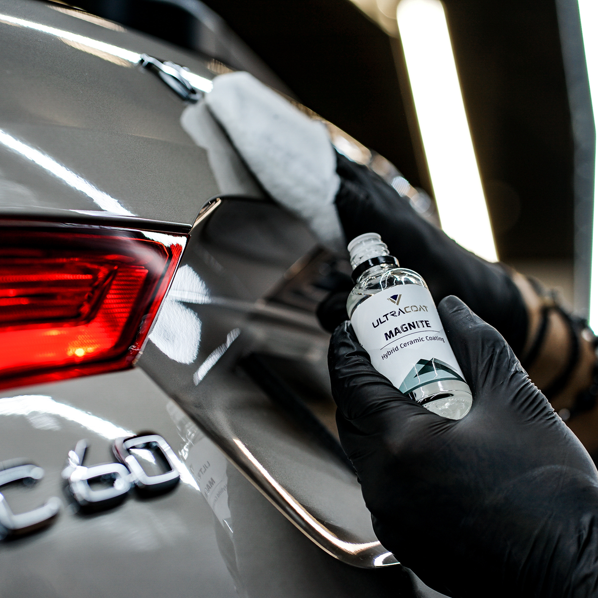 Gloved hand holding the Ultracoat Magnite bottle beside a dark vehicle's rear panel, with a microfibre cloth working the paintwork